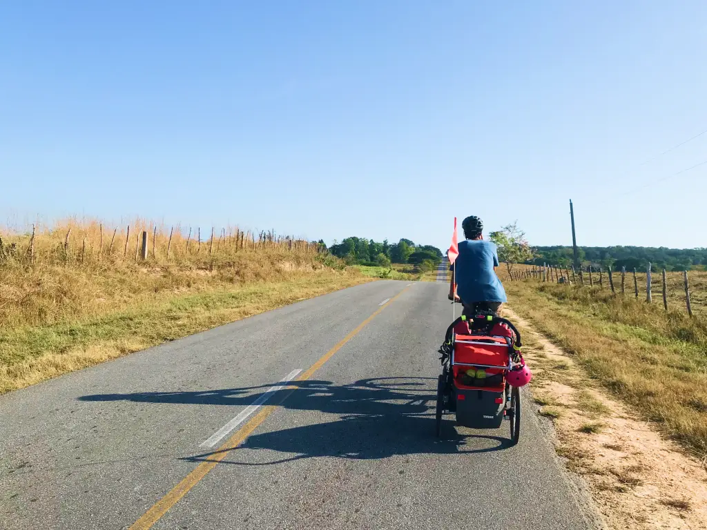 Cyclist towing a child in a trailer along a quiet countryside road in Cuba